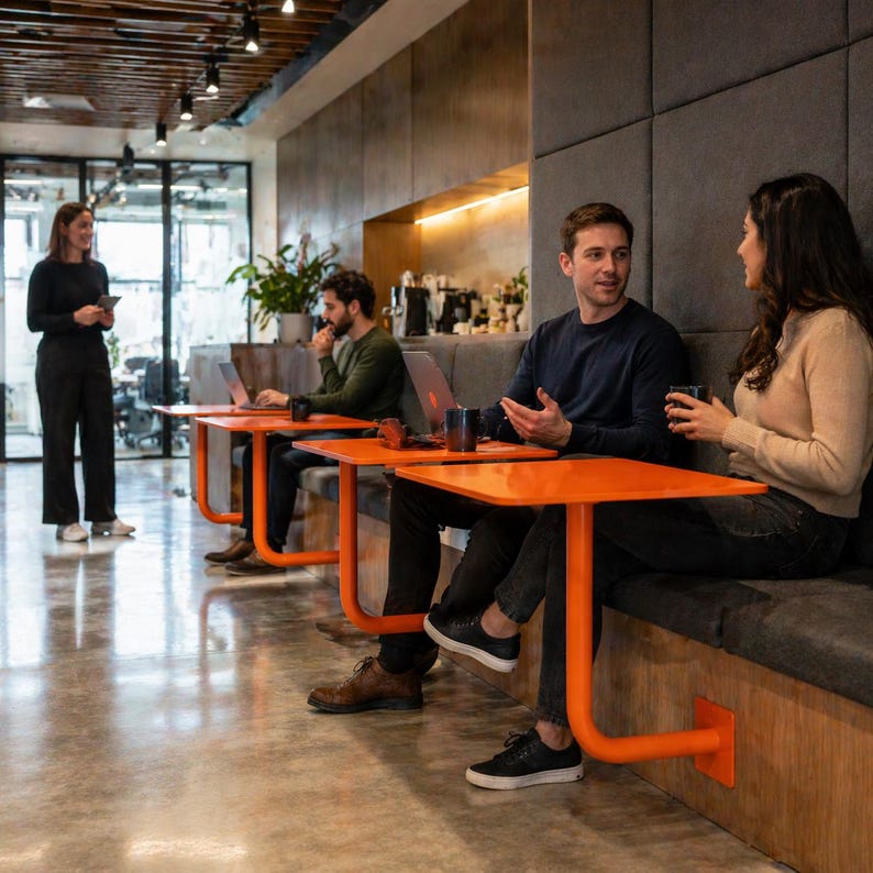 May include: A modern office space with several people interacting. Bright orange tables with curved legs are paired with built-in seating. One person stands holding a tablet, while others sit at tables, some with laptops and mugs.