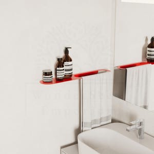May include: A contemporary red bathroom shelf with a towel rail. The shelf displays three brown bottles and a small jar. A white towel hangs from the rail. The shelf is mounted on a white wall, with a white sink and mirror in the background.