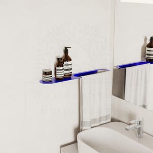May include: A contemporary blue bathroom shelf with brown bottles and a small jar. A white towel hangs beneath. The shelf is fixed to a white wall, with a similar shelf and mirror in view.