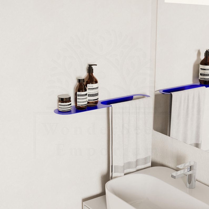 May include: A contemporary blue bathroom shelf with brown bottles and a small jar. A white towel hangs beneath. The shelf is fixed to a white wall, with a similar shelf and mirror in view.
