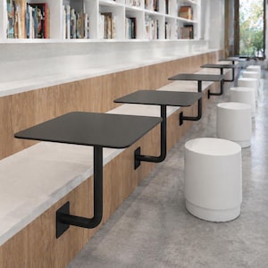 May include: A row of black square tables attached to a wooden bench, with white cylindrical stools. The tables are supported by black metal brackets. The background features a white bookshelf filled with books.
