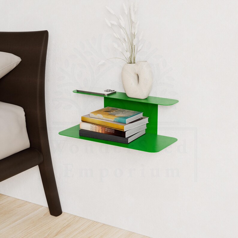 May include: A modern, bright green wall-mounted shelf with a minimalist design. The shelf holds a stack of books, a smartphone, and a white vase with decorative stems. The shelf is mounted on a white wall.