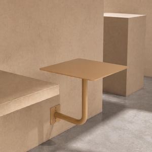 May include: A square, wall-mounted table in a muted tan color. The table has a single, curved support arm and is attached to a wall with a metal bracket. The design is minimalist and modern, suitable for small spaces.