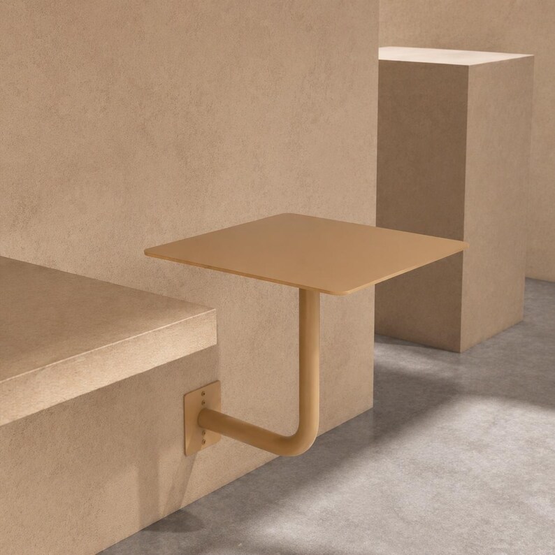 May include: A square, wall-mounted table in a muted tan color. The table has a single, curved support arm and is attached to a wall with a metal bracket. The design is minimalist and modern, suitable for small spaces.