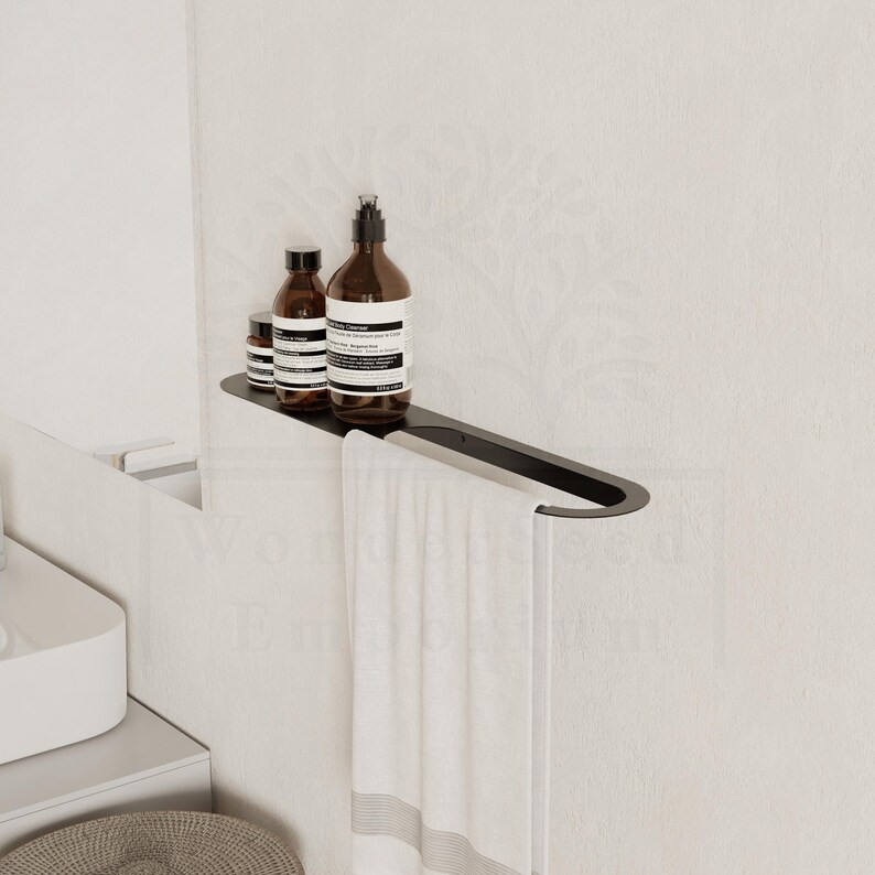 May include: A black metal shelf with a towel rail, holding three brown bottles. A white towel with a grey stripe hangs from the rail. The shelf is mounted on a white wall, creating a minimalist bathroom aesthetic.