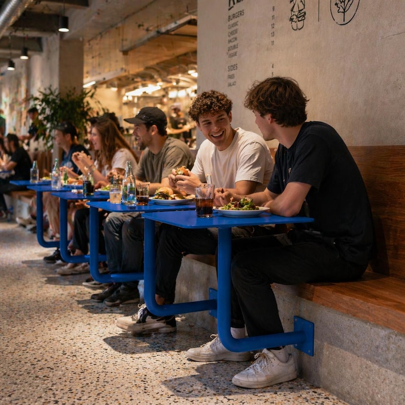 May include: A group of people seated at blue tables enjoying a meal in a restaurant. The tables are attached to a wooden bench and concrete wall. The people are eating burgers and salads. The restaurant has a modern design.