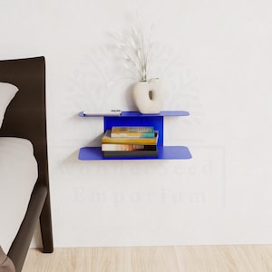 May include: A modern, cobalt blue wall-mounted shelf with two tiers. The top shelf holds a small white vase with decorative stems and a small, rectangular object. The lower shelf displays a stack of books. The shelf is mounted on a white wall.