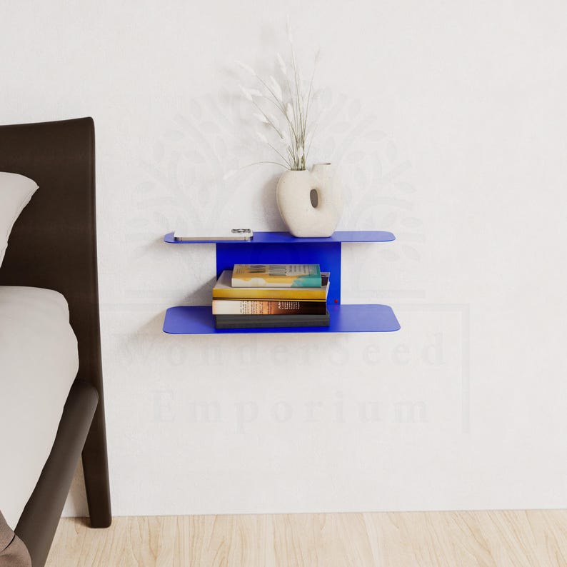 May include: A modern, cobalt blue wall-mounted shelf with two tiers. The top shelf holds a small white vase with decorative stems and a small, rectangular object. The lower shelf displays a stack of books. The shelf is mounted on a white wall.