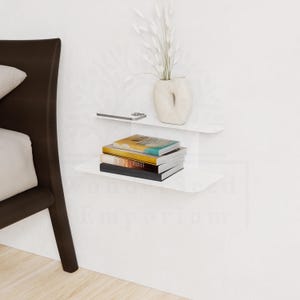May include: White floating shelves mounted on a white wall, holding a stack of books, a smartphone, and a decorative vase with white dried flowers. The shelves are curved and minimalist in design.