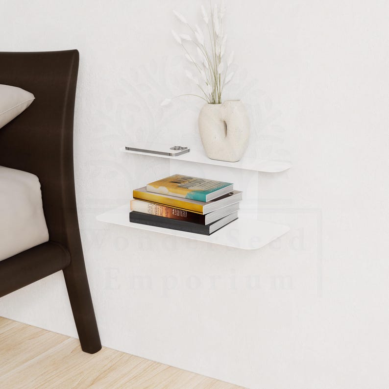 May include: White floating shelves mounted on a white wall, holding a stack of books, a smartphone, and a decorative vase with white dried flowers. The shelves are curved and minimalist in design.