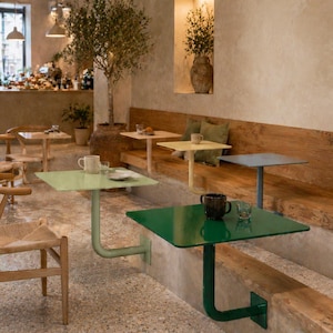 May include: A cafe interior featuring several small, square tables in shades of green, yellow, and blue. Each table is attached to the wall with a curved metal support. The tables are set with mugs and small plates, suggesting a dining area.