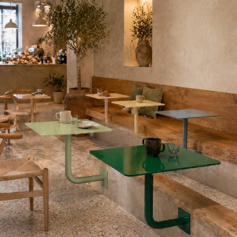 May include: A cafe interior featuring several small, square tables in shades of green, yellow, and blue. Each table is attached to the wall with a curved metal support. The tables are set with mugs and small plates, suggesting a dining area.