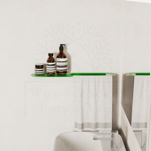 May include: A contemporary bathroom scene with a green shelf displaying three brown bottles with white labels. Below, a green towel rail holds a white towel. The backdrop is a white wall with a delicate floral design.