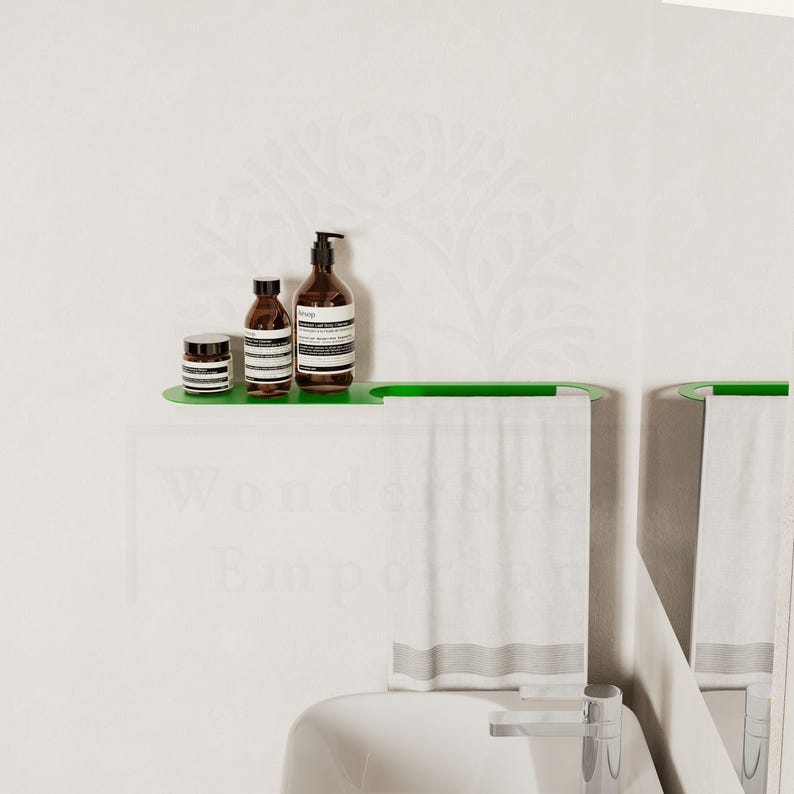 May include: A contemporary bathroom scene with a green shelf displaying three brown bottles with white labels. Below, a green towel rail holds a white towel. The backdrop is a white wall with a delicate floral design.