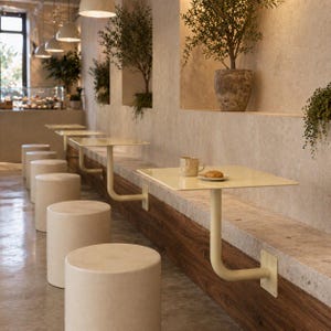 May include: A cafe interior featuring a row of cream-colored cylindrical stools and square tables. The tables are attached to a wooden and stone wall. Olive trees in pots and hanging plants add greenery. A cup and a pastry sit on one table.