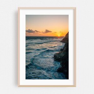 Könnte beinhalten: Ein gerahmter Druck eines Sonnenuntergangs an der Küste. Das Bild zeigt die Sonne, die über dem Meer untergeht, mit Wellen, die gegen eine felsige Klippe schlagen. Der Himmel wechselt von Orange zu Blau, mit ein paar Wolken.
