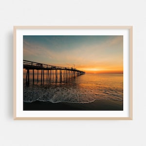 May include: A framed landscape photograph of a wooden pier extending into the ocean at sunrise. The sky transitions from deep blue to vibrant orange, reflecting on the calm water surface. Gentle waves wash onto the dark sandy shore below.
