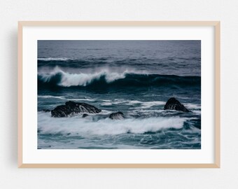 Op de afbeelding: Een ingelijste print met een zeegezicht met donkerblauw water en witte golven die op donkere rotsen breken. De afbeelding toont de kracht en beweging van de oceaan, met een stemmingvolle, atmosferische uitstraling. De lijst is licht van kleur.
