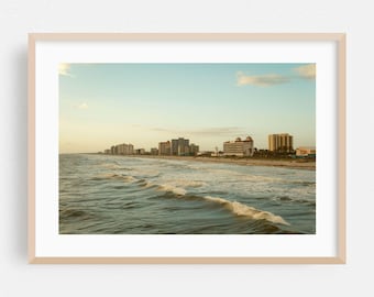 May include: A framed print of a coastal scene. The image captures a view of the ocean with waves rolling towards the shore. Buildings line the beach under a blue sky with a few clouds. The overall color palette is warm, with golden and blue tones.