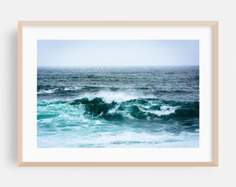Op de afbeelding: Een ingelijste prent van de oceaan met turquoise en blauwe golven. Het water is woelig, met wit schuim zichtbaar als de golven breken. De lucht is lichtblauw en de prent zit in een licht houten frame.