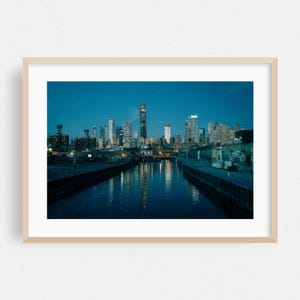 以下が含まれることがあります： 夕暮れ時の都市景観の額入りプリント。画像は、都市の光を反射する両側に建物のある川または運河を捉えています。空は深い青色で、建物は照らされています。