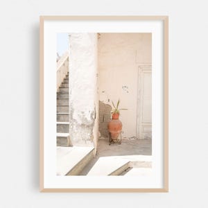 May include: A framed wall art print featuring a light-colored building exterior with a stone staircase, a white door, and a terracotta-colored clay pot on a metal stand containing a green plant, all set against a weathered, textured wall.