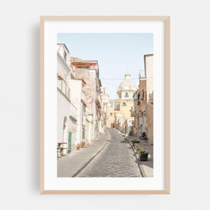 May include: A framed art print featuring a narrow, sunlit cobblestone street in an Italian town. The scene includes light-colored buildings with balconies, potted plants along the path, and a prominent domed church building in the distance.