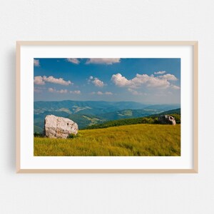 May include: A framed landscape photograph featuring a vibrant mountain scene. The image showcases a bright blue sky dotted with fluffy white clouds, rolling green hills, and two large, light-colored boulders in a field of golden grass.