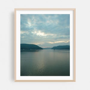 May include: A framed landscape print showing a calm lake scene beneath a cloudy sky. The water mirrors the sky's hues, with green hills on either side. The print is in a light-coloured wooden frame.