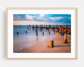May include: A framed photograph of a beach scene at sunset. The image features weathered wooden pilings and remnants of a pier extending into calm water. The sky is a mix of blue and orange hues.