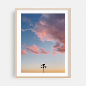 May include: Framed print of a sunset sky with pink and purple clouds against a blue background. A palm tree silhouette is at the bottom, set against an orange and yellow gradient. The image is framed in a light wood.