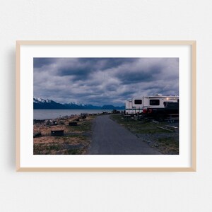 May include: A framed landscape photograph showing a paved path leading toward a body of water under a dark, cloudy sky. White recreational vehicles are parked on the right side, and snow-capped mountains are visible in the distant background.