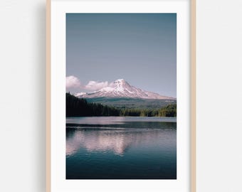 May include: A framed vertical landscape photograph featuring a snow-capped mountain peak rising above a dark forest and a calm lake. The mountain is reflected in the water, and the sky is a clear, muted blue. The frame is a light-toned wood.