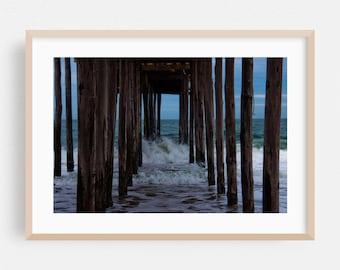 May include: A framed photograph of a pier with dark brown wooden pilings. The ocean waves crash beneath the pier, with a blue sky visible in the distance. The image is framed in a light beige wooden frame.