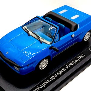May include: A bright blue scale model of a 1987 Lamborghini Jalpa Spyder Prototipo convertible car. The miniature vehicle features a black interior, silver wheels, and windscreen wipers. The base is labelled Lamborghini Jalpa Spyder (Prototipo) (1987) 1 of 1.000 pcs.
