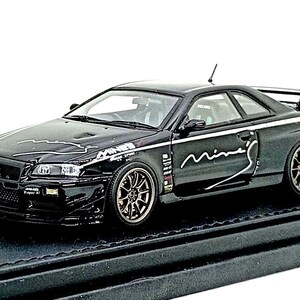 Könnte beinhalten: Ein schwarzes Modellauto des Nissan Skyline GT-R R34 Coupés mit bronzefarbenen Mehrspeichenfelgen, einem Heckspoiler und weißen Mine's-Aufklebern auf der Motorhaube und den Seiten. Das Auto ist auf einem schwarzen Sockel mit silbernem Schild montiert.