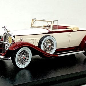 Può includere: Un modellino in scala di un'auto decappottabile d'epoca con una verniciatura bicolore color crema e rosso scuro. Il veicolo presenta ruote a raggi cromate con pneumatici a fascia bianca, una ruota di scorta laterale e fari gialli.