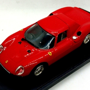 May include: A red die-cast model sports car featuring a low-profile aerodynamic body, a black windscreen, and silver-toned wheels. The vehicle displays small yellow Ferrari emblems on the bonnet and front wings, with a black interior visible.