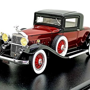 Op de afbeelding: Gedetailleerd model van een klassieke bordeauxrode en zwarte coupé. De auto heeft whitewall-banden met rode velgen, chromen accenten en een zwart dak. De voorkant heeft een chromen grille en koplampen.