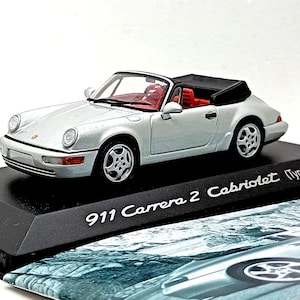 May include: A silver Porsche 911 Carrera 2 Cabriolet (Typ 964) model car with a black convertible top and red interior. The model sits on a black base with the car's name printed on it.