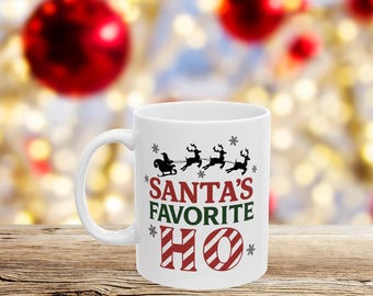 May include: White ceramic mug with the text "SANTA'S FAVORITE HO" in red and green, with a black silhouette of Santa's sleigh and reindeer. Snowflakes are scattered around the text. The mug is on a wooden surface with a blurred Christmas background.