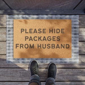 May include: A rectangular doormat with the text "PLEASE HIDE PACKAGES FROM HUSBAND" in dark gray letters. The mat has a tan center and a gray and white checkered border. Black sneakers and blue jeans are visible.