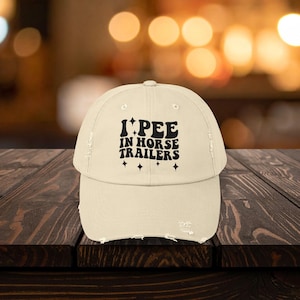 May include: A distressed, cream-colored baseball cap with the text "I PEE IN HORSE TRAILERS" in black, retro-style lettering. Four small black stars are below the text. The cap has a curved brim and a worn appearance.
