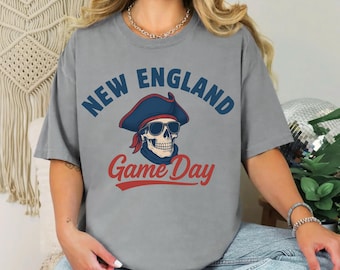 May include: Gray t-shirt with the words "NEW ENGLAND" in blue and "Game Day" in red. A skull wearing a tricorn hat, sunglasses, and a red scarf is in the center. The shirt is a casual style.