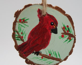 May include: A hand-painted wooden ornament featuring a red cardinal perched on a branch with green leaves and red berries. The ornament is painted on a round piece of wood with a natural bark edge.