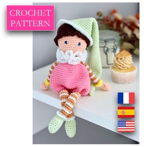 Puede incluir: Patrón de crochet para una muñeca rosa y marrón con un gorro de dormir verde. La muñeca está sentada en un estante blanco con un cupcake en el fondo. El patrón está disponible en francés, español e inglés.