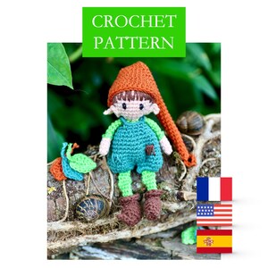 Może przedstawiać: Wzór na szydełku dla małego gnoma z pomarańczowym kapeluszem, niebieskimi ogrodniczkami i zielonymi nogami. Gnom siedzi na kłodzie z zielonymi liśćmi i ślimakami. Wzór jest dostępny w języku francuskim, angielskim i hiszpańskim.