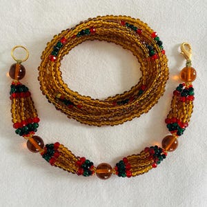 May include: A golden beaded waist chain with red and green accents. The chain is made of small, round beads and features decorative tassels with larger amber-colored beads. The chain is coiled and laid out on a white surface.