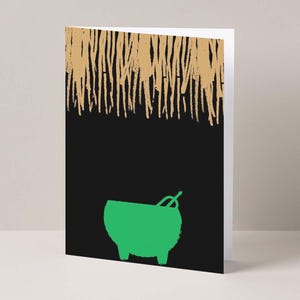 May include: Greeting card with a black background and a green cauldron illustration. The top of the card features a gold, textured design. The card is standing upright on a white surface.