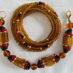 May include: An amber and gold beaded waist chain with red and green accents. The chain is made of small, round beads and has decorative sections with larger beads and gold details. Designed to be worn around the waist.